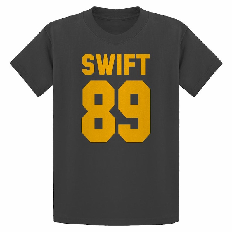 May include: A dark gray t-shirt with the word "SWIFT" in gold block letters above the number "89" in gold. The shirt is a crew neck style and appears to be made of a soft material.
