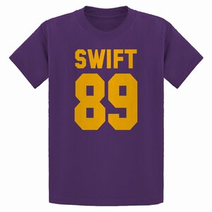 May include: Purple t-shirt with the name "SWIFT" and the number "89" printed in gold. The text and numbers are in a bold, blocky font. The shirt has short sleeves and a crew neck.