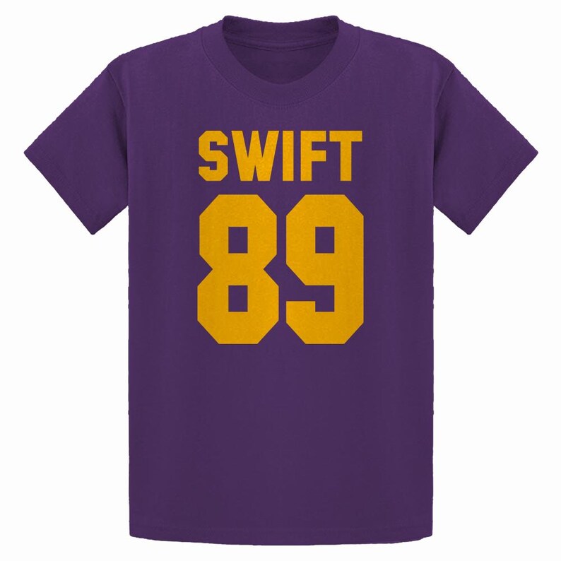 May include: Purple t-shirt with the name "SWIFT" and the number "89" printed in gold. The text and numbers are in a bold, blocky font. The shirt has short sleeves and a crew neck.