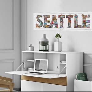 May include: A white desk with a fold-down top, a laptop computer, and a vase with a plant on the top shelf. The desk is against a white wall with a framed print of the word "SEATTLE" made of images of the city.