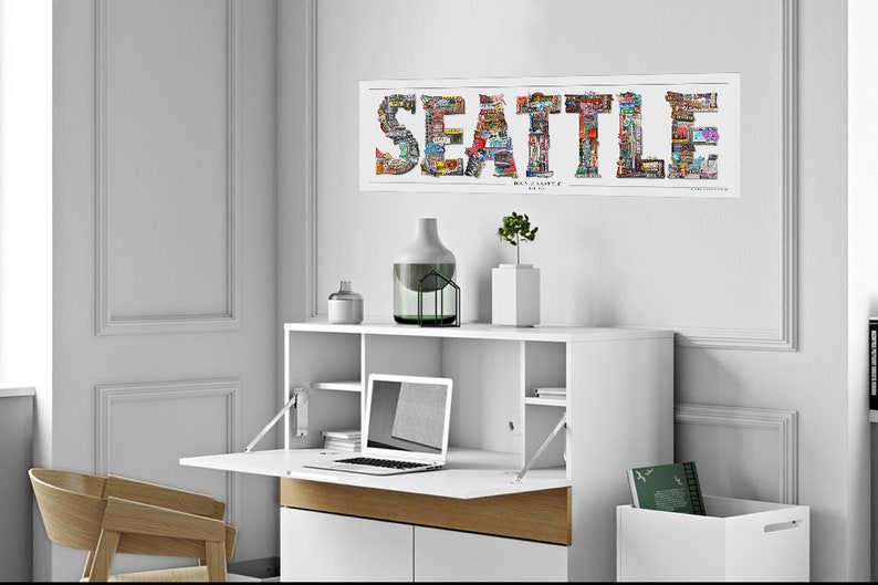May include: A white desk with a fold-down top, a laptop computer, and a vase with a plant on the top shelf. The desk is against a white wall with a framed print of the word "SEATTLE" made of images of the city.