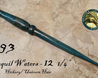 Op de afbeelding: Een groenblauwe houten toverstaf met een taps toelopend handvat en een gladde afwerking. De tekst #793, Tranquil Waters - 12 1/4", Hickory/Unicorn Hair is zichtbaar. Een rond logo toont een drakenkop met de tekst Dragon Alley Wands.