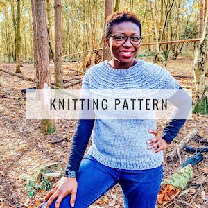 May include: A light blue and white knitted sweater with the text "KNITTING PATTERN" overlaid on the image. The sweater has a textured collar and is worn with a long-sleeved black shirt and blue jeans.