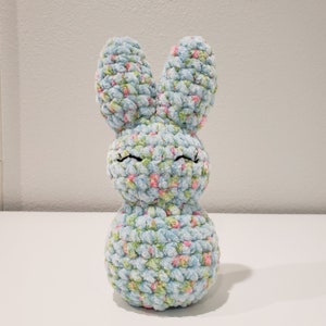 May include: A light blue crocheted bunny with pink and green floral accents. The bunny has black embroidered eyes and a smiling expression.