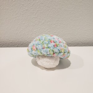 May include: A blue and pink crocheted mushroom with a white stem. The mushroom is made of soft yarn and has a floral pattern.