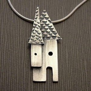 May include: A silver pendant necklace featuring a pair of stylized houses with textured roofs. The houses have a simple, geometric design with rounded tops and small windows.