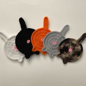 May include: A set of five circular crochet coasters shaped like animal backsides, each featuring a small upright tail and a centered pink stitched detail. The coasters are crafted in white, black, orange, light gray, and a variegated brown yarn.
