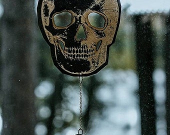 Op de afbeelding: Een zwarte en gouden schedelvormige ornament hangt aan een gouden ketting met een halve maan en een kristal hanger. De ornament hangt voor een wazig raam.