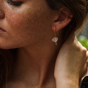 May include: A close-up of a person's ear with a gold earring featuring a small, white, irregular-shaped stone.