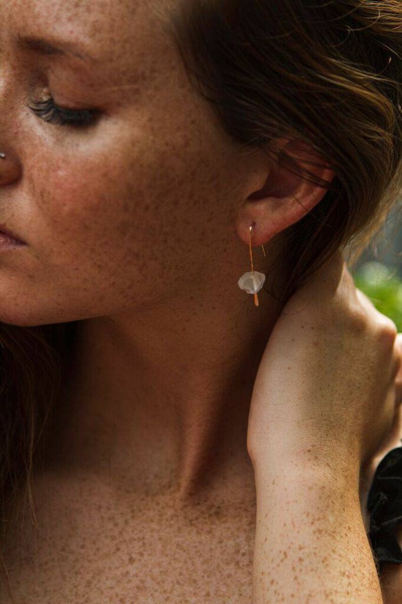 May include: A close-up of a person's ear with a gold earring featuring a small, white, irregular-shaped stone.