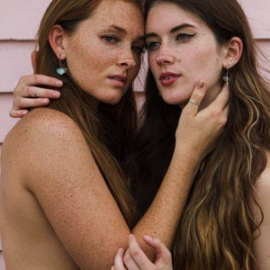 May include: Two women with long hair, one with freckles, are standing close together. They are both looking at the camera. The woman on the left is wearing a teal blue top. The woman on the right is wearing no top. They are both wearing earrings.