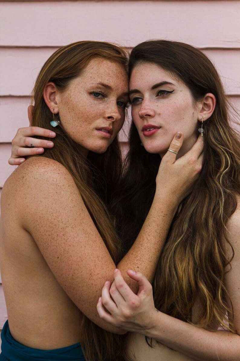 May include: Two women with long hair, one with freckles, are standing close together. They are both looking at the camera. The woman on the left is wearing a teal blue top. The woman on the right is wearing no top. They are both wearing earrings.