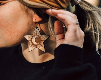 May include: A pair of wooden star-shaped earrings with a crescent moon and sun design. The earrings are a light brown color with gold accents.