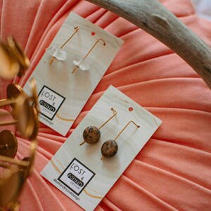 May include: Two pairs of earrings on white cards with the text "Lost & Found Design" and the social media handle "@lostandfounddesign". The earrings on the left are made of clear quartz and gold wire. The earrings on the right are made of brown and black jasper and gold wire.