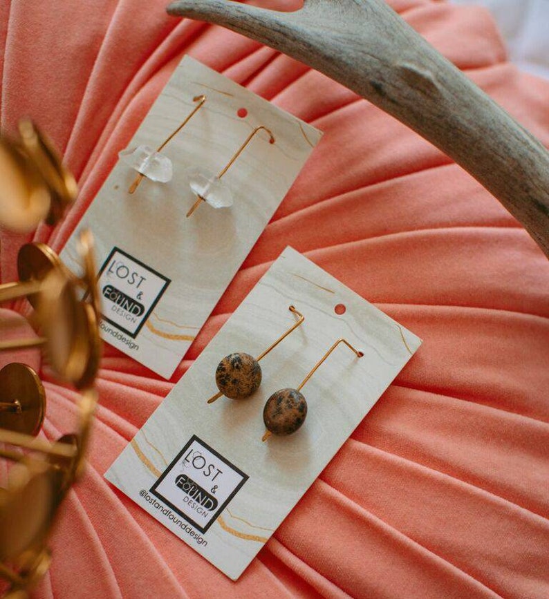 May include: Two pairs of earrings on white cards with the text "Lost & Found Design" and the social media handle "@lostandfounddesign". The earrings on the left are made of clear quartz and gold wire. The earrings on the right are made of brown and black jasper and gold wire.