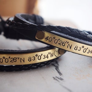 May include: Two black leather bracelets with braided straps and gold metal plates engraved with the coordinates "40°0′26″N 83°0′34″W".