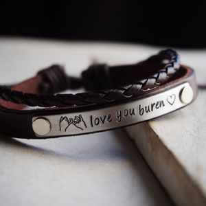 May include: Brown leather bracelet with a silver plate engraved with the text "I love you buren" and a small heart symbol. The plate also features a small graphic of two hands making a pinky promise.