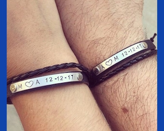 May include: Two black braided leather bracelets featuring a rectangular silver-toned metal plate. One plate is engraved with M, a heart symbol, A, and the date 12-12-17. The other plate is engraved with A, a heart symbol, M, and the date 12-12-17.