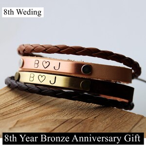 Pode incluir: Uma pilha de pulseiras com duas tiras de couro trançado em castanho e castanho escuro, e duas tiras de metal planas em tons de cobre e latão. As tiras de metal estão gravadas com as letras B, um símbolo de coração e J. Texto: 8th Weding.