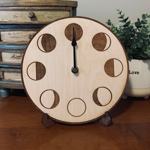 May include: A wooden clock with a moon phase design. The clock face is light brown with dark brown moon phases. The clock hands are black and the clock is standing on a wooden surface.