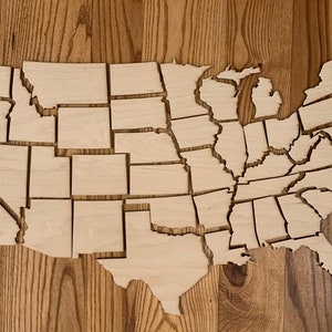 May include: A wooden puzzle of the United States of America, with each state cut out individually. The puzzle pieces are arranged in the shape of the country, with Alaska and Hawaii off to the side.
