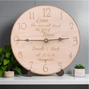 May include: A round wooden clock with a light brown face and dark brown hands. The clock face features the text "Love like ours will stand the test of Time" and the names "Danielle & Mark" with the date "est. 12/5/14". The clock is displayed on a stand.