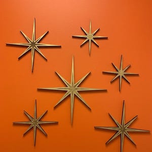 May include: A collection of six wooden starburst wall decorations in varying sizes. Each star features a multi-pointed design with sharp, elongated rays radiating from a central point, showcasing a natural wood grain finish with dark edges.