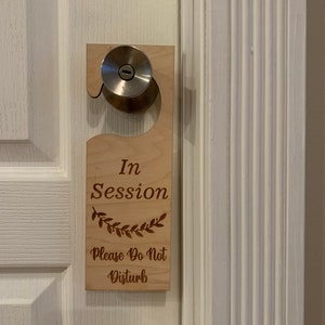 May include: A wooden door hanger with the text "In Session Please Do Not Disturb" and a leafy design. The hanger is attached to a door knob.