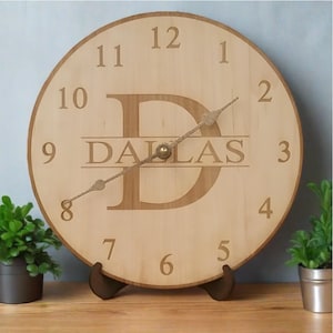 May include: A wooden clock with a large letter "D" in the center, with the word "DALLAS" written below it. The clock face has Roman numerals and a gold-colored hand.