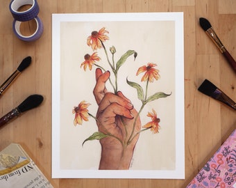 以下が含まれることがあります： オレンジと黄色の花を持つ手の水彩画プリント。アートワークは白い背景にあります。筆、テープ、花柄のノートが、木製の表面に印刷物の周りに配置されています。