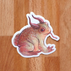 May include: A sticker featuring a whimsical creature with a frog-like body, horns, and a long, red tongue. The sticker has a white border and is primarily shades of brown, red, and green. The creature is in a seated position.