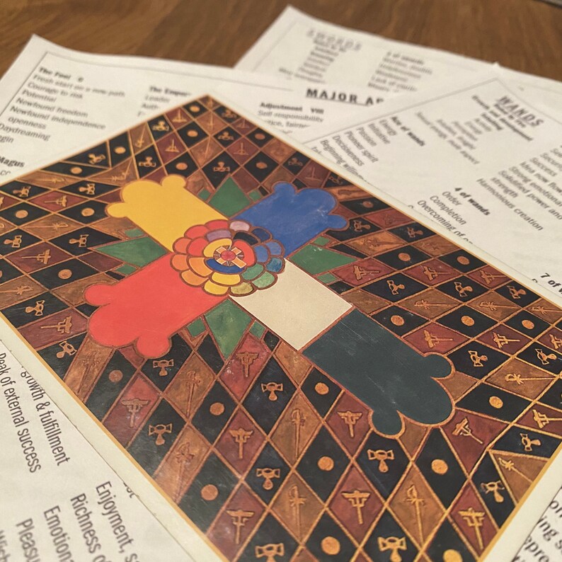 K&ouml;nnte beinhalten: Eine Tarotkarte mit einem farbenfrohen Design, das ein Kreuz mit vier Farben zeigt, rot, gelb, blau und gr&uuml;n. Die Karte ist vor einem Hintergrund aus einem sich wiederholenden Muster aus schwarzen und goldenen Rauten platziert. Die Karte ist mit "7 of Wands" beschriftet und hat den Text "Peak of external success, Growth & fulfillment" in der linken unteren Ecke.