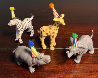 May include: Four small plastic animal figurines including a zebra, a giraffe, a hippopotamus, and an elephant. Each animal wears a miniature, colorful striped party hat topped with a small, fuzzy pom-pom in yellow, orange, blue, or green.