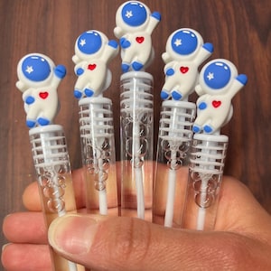 May include: A collection of bubble wands featuring astronaut figurines. Each wand has a clear plastic tube filled with bubble solution, topped with a white astronaut figure with blue accents and a red heart. The wands are held in a hand.