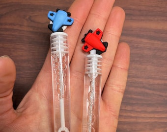 May include: Two clear plastic bubble wands filled with liquid. One wand features a light blue airplane topper, and the other features a red airplane topper. Each wand has a white plastic internal wand piece with a circular loop at the bottom.