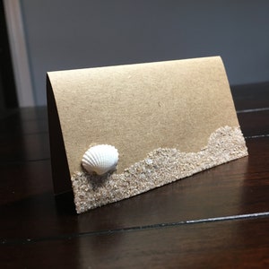 May include: A brown card with a white seashell and a border of sand glued to the bottom.