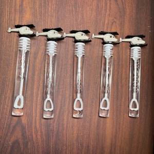 May include: Five clear plastic bubble wands arranged in a row. Each wand features a white plastic helicopter topper with black rotor blades. The wands have a long, transparent cylindrical tube containing a white bubble wand applicator inside.
