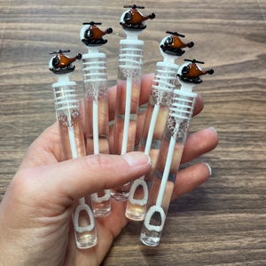 May include: Five clear plastic bubble wands, each topped with a small brown and black helicopter figurine. The wands feature a white internal stem with a circular loop at the bottom and a white cap that secures the helicopter topper to the tube.