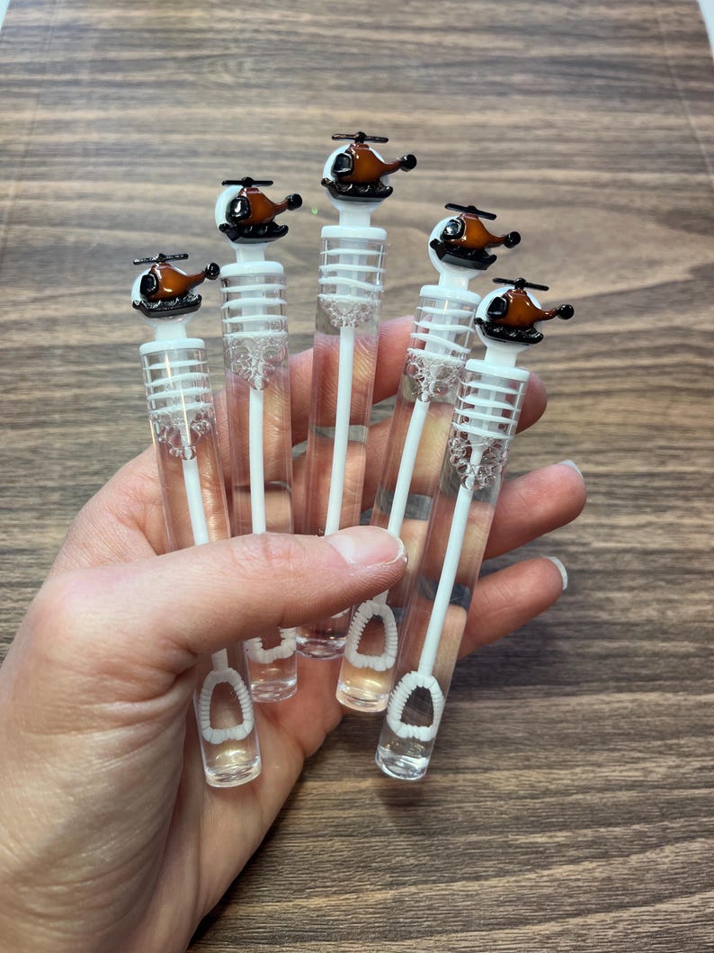 May include: Five clear plastic bubble wands, each topped with a small brown and black helicopter figurine. The wands feature a white internal stem with a circular loop at the bottom and a white cap that secures the helicopter topper to the tube.