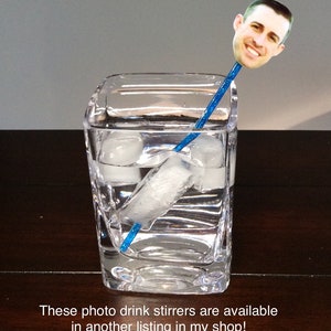Puede incluir: Un agitador de bebidas de pl&aacute;stico azul con una foto de una persona en la parte superior. El agitador est&aacute; en un vaso de agua con cubitos de hielo.
