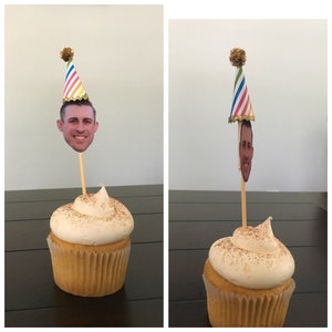 Puede incluir: Dos cupcakes con glaseado blanco y una pizca de canela encima. Cada cupcake tiene un gorro de fiesta con una cara en la parte superior. Los gorros de fiesta son de rayas arco&iacute;ris con ribetes dorados y un pomp&oacute;n dorado en la parte superior.