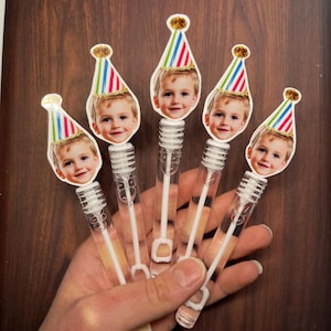 May include: Five bubble wands with a child's face and a party hat attached. The wands are clear plastic tubes with a bubble solution inside. The party hats are red, blue, and green striped. The background is a dark wood surface.