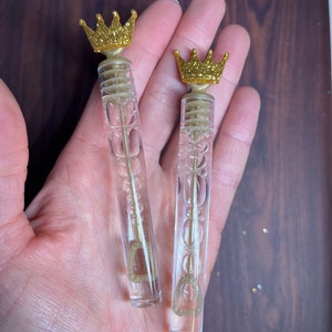 May include: Two clear plastic bubble wands with gold glitter crowns on top. The wands are held in a hand.