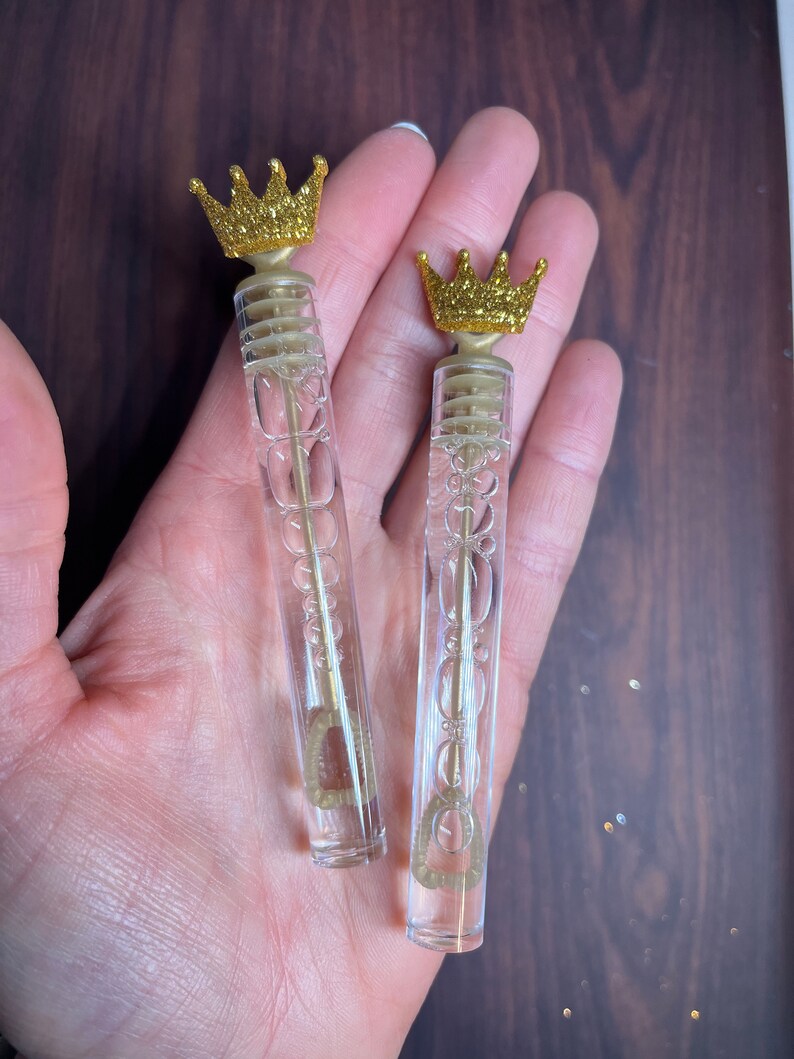 May include: Two clear plastic bubble wands with gold glitter crowns on top. The wands are held in a hand.