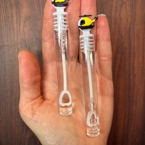 May include: Two clear plastic bubble wands featuring yellow and black helicopter toppers. Each wand consists of a transparent tube filled with bubble solution, a white internal wand mechanism, and a detailed helicopter-shaped cap in yellow and black.