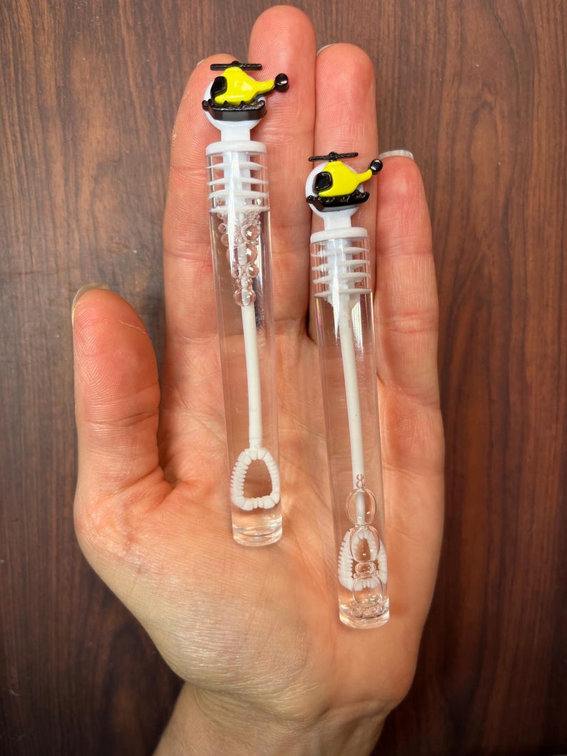 May include: Two clear plastic bubble wands featuring yellow and black helicopter toppers. Each wand consists of a transparent tube filled with bubble solution, a white internal wand mechanism, and a detailed helicopter-shaped cap in yellow and black.