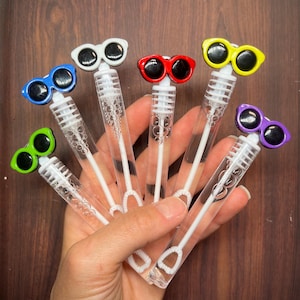May include: A collection of six clear plastic bubble wands, each topped with a pair of miniature sunglasses in different colors including blue, white, red, yellow, green, and purple. Each wand contains a white plastic bubble blowing tool inside.