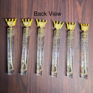 May include: Six gold and clear plastic bubble wands with a crown and heart design on the top. The wands are arranged in a row on a brown wooden surface. Back View.