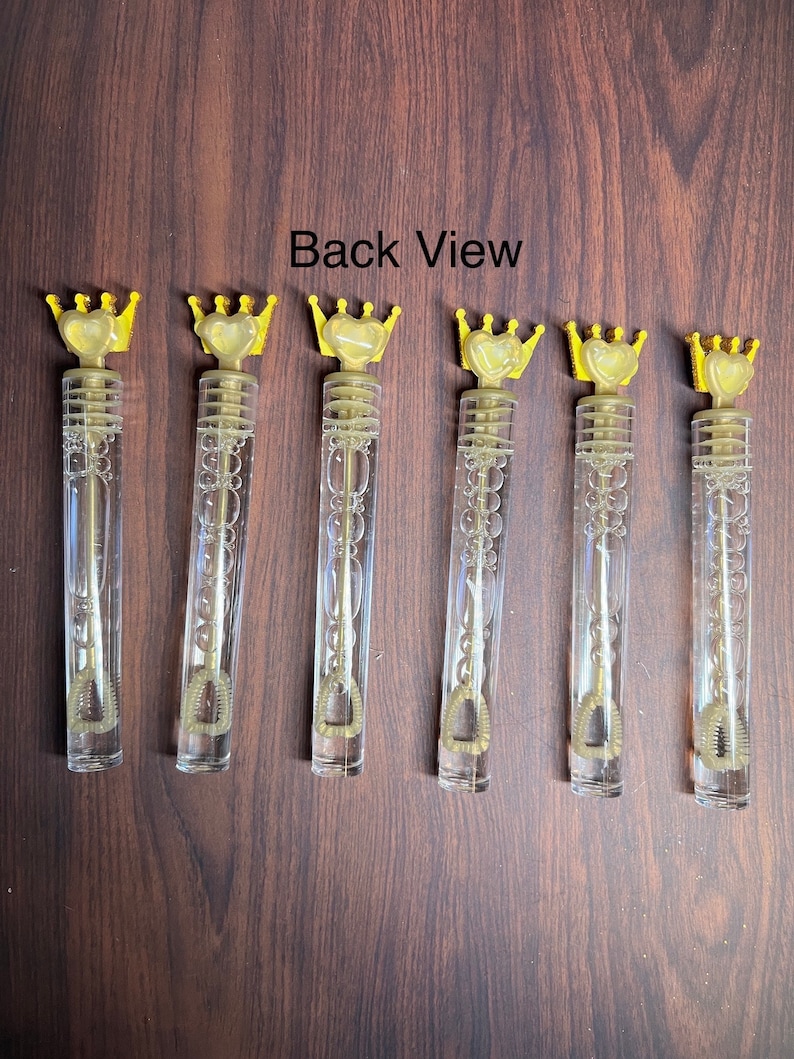 May include: Six gold and clear plastic bubble wands with a crown and heart design on the top. The wands are arranged in a row on a brown wooden surface. Back View.