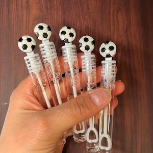 May include: Five clear plastic bubble wands, each topped with a small black and white soccer ball figurine. The wands contain a transparent liquid and feature a white internal wand mechanism with a circular loop at the bottom for blowing bubbles.